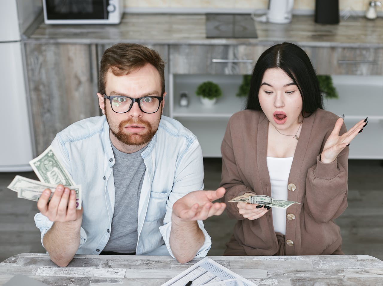 Interracial couple shocked while handling cash in a modern kitchen. Captivating expressions indoors.