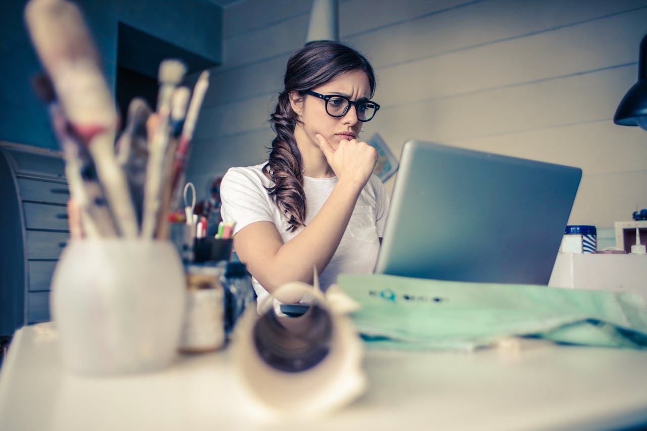 project-02-c Young woman with glasses deeply focused on a laptop surrounded by art supplies in a home office.